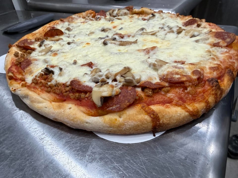 A freshly baked pizza with pepperoni, sausage, mushrooms, and melted cheese on a metal counter.